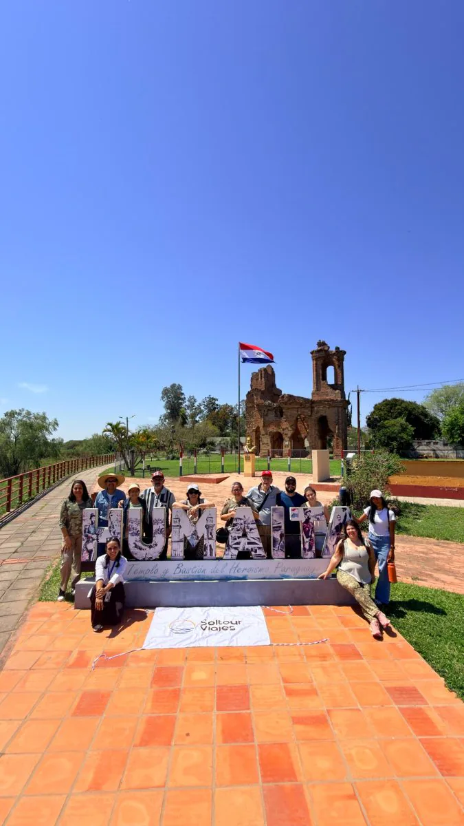 Grupo de viajeros de Soltour junto al cartel de Humaitá, con las ruinas de la iglesia y la bandera paraguaya detrás